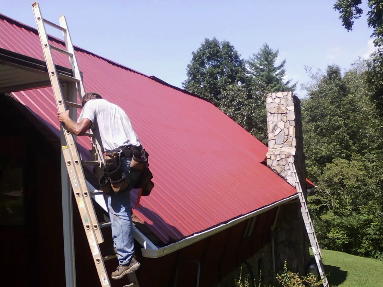 Roofer on a ladder beside a red metal roof and stone chimney — Soffit & Fascia Repair by Roofing Brothers in Easton